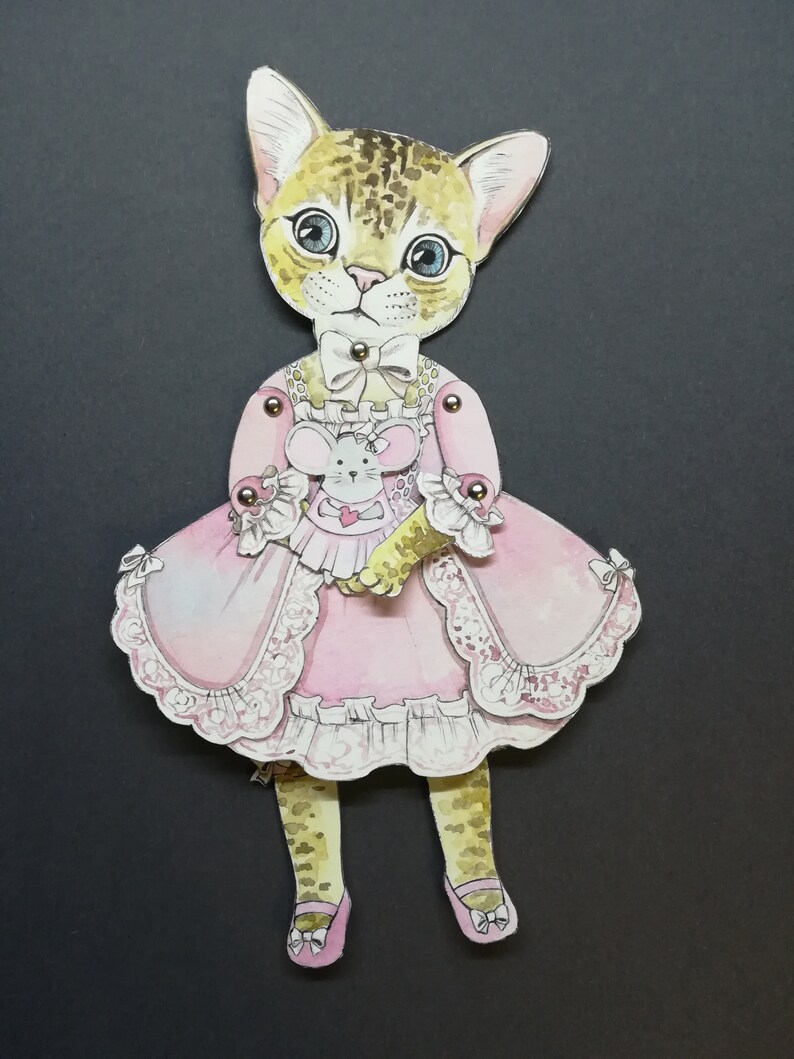 May include: A paper doll of a cat wearing a pink dress with lace trim and holding a small mouse doll. The cat has brown and black spots and blue eyes.