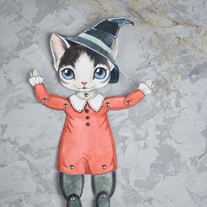 May include: A paper doll of a cat wearing a witch's hat and a red dress. The doll has movable arms and legs.
