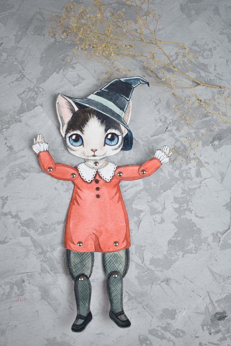May include: A paper doll of a cat wearing a witch's hat and a red dress. The doll has movable arms and legs.