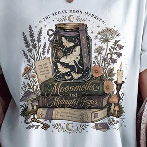 Könnte beinhalten: Ein weißes T-Shirt mit einem Grafikdruck eines mit Motten gefüllten Glasgefäßes, das auf einem Stapel aus drei Büchern ruht. Der Text lautet THE SUGAR MOON MARKET und Moonmoths And Midnight Pages. Das Design enthält botanische Elemente, einen Schlüssel und eine Kerze.