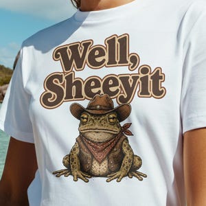 Peut inclure: T-shirt blanc avec l'inscription "Well, Sheeeyit" en lettres marron. Le t-shirt présente une grenouille de dessin animé portant un chapeau de cow-boy et un bandana. L'arrière-plan montre une scène de plage tropicale avec de l'eau bleue et un palmier.