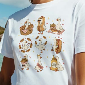 May include: A white short-sleeved t-shirt featuring a graphic print of nine cartoon ducks in various poses, including a duck in a picnic basket, a duck dressed as a hot dog, and a duck holding a bottle labeled Yellow Mustard.