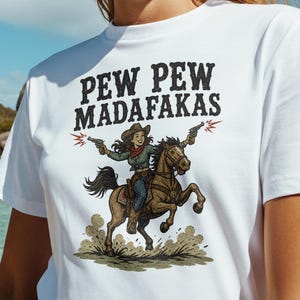 以下が含まれることがあります： 「PEW PEW MADAF*KAS」の文字と、馬に乗って銃を撃つ人のグラフィックが描かれた白いTシャツ。その人はカウボーイハットをかぶっています。背景にはビーチ、海、ヤシの木があります。
