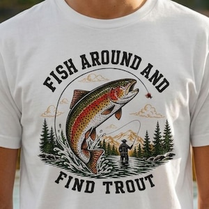 May include: White t-shirt with a graphic of a trout jumping out of water, a fisherman, and the text "Fish Around and Find Trout". The design includes mountains and trees. The shirt is worn by a person with curly hair.