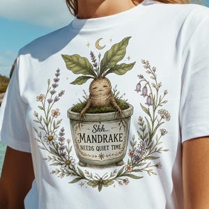 May include: A white t-shirt featuring a graphic of a mandrake plant in a pot with the text Shh... MANDRAKE NEEDS QUIET TIME, surrounded by a floral wreath illustration. The design includes a crescent moon and small moth at the base.