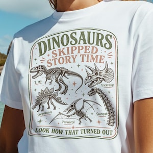May include: White t-shirt with a dinosaur graphic. The design features dinosaur skeletons and illustrations with the text "DINOSAURS SKIPPED STORY TIME" and "LOOK HOW THAT TURNED OUT". The shirt is worn outdoors with a tropical background.
