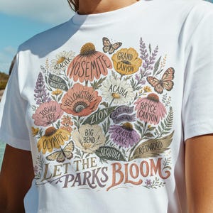 May include: White t-shirt with a floral design featuring national park names like Yosemite and Grand Canyon. The shirt includes the text "Let the Parks Bloom" in a decorative font. The image is taken outdoors with a tropical background.