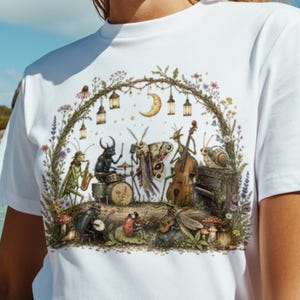 May include: A white short-sleeved t-shirt featuring a detailed graphic print of various insects playing musical instruments under a crescent moon and hanging lanterns, framed by a natural arch of vines, flowers, and mushrooms.