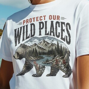 May include: White t-shirt with the text "PROTECT OUR WILD PLACES" above a graphic of a bear with a mountain and forest scene. The bear has a river running through it. The person is wearing pink sunglasses.