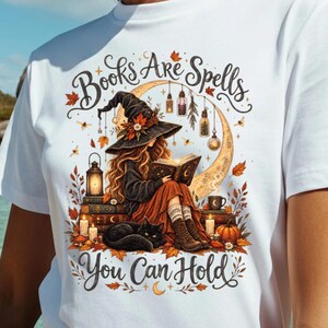 May include: A white t-shirt featuring a graphic illustration of a person in a witch hat reading a book, surrounded by autumn leaves, candles, and a black cat. The text reads "Books Are Spells You Can Hold" in a decorative script font.