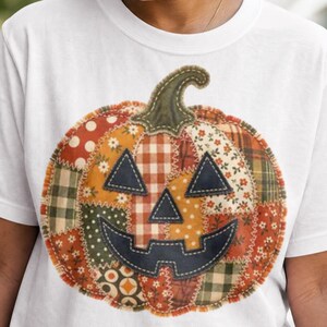 May include: A white short-sleeve t-shirt featuring a large, central graphic of a jack-o'-lantern pumpkin. The pumpkin is designed with a patchwork pattern of various autumn-colored fabrics, including orange, brown, and green plaids, florals, and polka dots, with dark navy blue felt-like eyes, nose, and a jagged mouth.