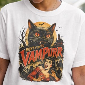 May include: White t-shirt featuring a vintage-style graphic. The design includes a black cat with orange eyes, a full moon, and the text "NIGHT OF THE VAMPURR". A woman in a red dress is also depicted, with a spooky graveyard scene.