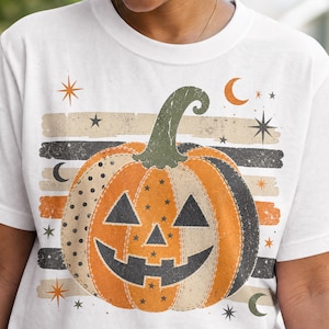 Puede incluir: Una camiseta blanca con un gráfico grande de calabaza de Halloween. La calabaza es naranja, beige y gris oscuro con patrones de puntos y estrellas. Está sobre rayas horizontales, estrellas y lunas crecientes con un estilo desgastado.