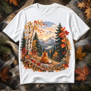 May include: A white short-sleeve t-shirt featuring a circular graphic print of a fox sitting on a rock, overlooking a mountain landscape with birch and pine trees, autumn foliage, and a sunset sky with birds flying in the distance.