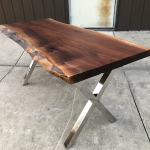 May include: A large, live edge walnut wood table top with a natural, brown wood finish. The table top is supported by a set of four silver, X-shaped legs.