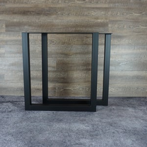 May include: Two black metal table legs with a rectangular frame. The legs are made of square tubing and have a simple, modern design.