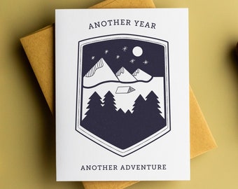 May include: A white card with a navy blue shield design featuring a mountain range, a small cabin, a moon, and stars. The text "Another Year Another Adventure" is printed on the card.