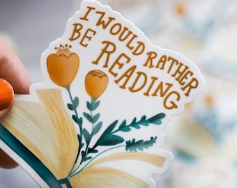 Può includere: Un adesivo bianco con il testo "I would rather be reading" in lettere marroni. L'adesivo presenta un'illustrazione stilizzata di un libro aperto con fiori gialli e foglie verdi.