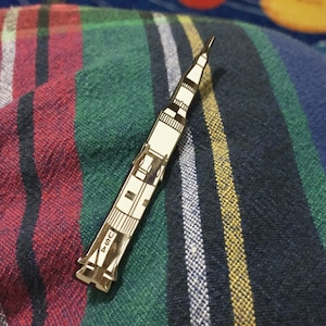 May include: A silver-coloured pin shaped like a rocket ship with the word "USA" on the side. The pin is on a striped fabric background.