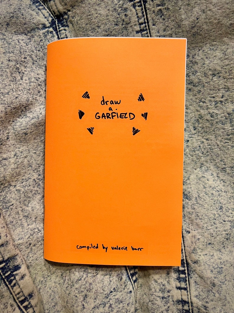 May include: An orange booklet with the text 'draw a GARFIELD' in black marker. The booklet has a black marker drawing of a circle with arrows around it. The bottom of the booklet has the text 'compiled by valerie barr' in black marker.