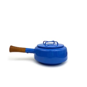 Puede incluir: Una olla de fondue de esmalte azul con tapa y mango de madera. La olla tiene un cuerpo redondo y un mango largo. La tapa tiene un asa metálica. La olla está diseñada para cocinar y servir fondue.