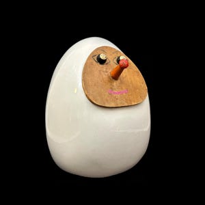 May include: A whimsical, white egg-shaped figurine with a wooden face. The face features painted black eyes, a red-tipped orange nose, and a pink smiling mouth. The figurine is set against a black background.