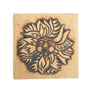 May include: A square ceramic tile featuring a brown and black sun design. The sun has a face with a wide smile and large eyes.