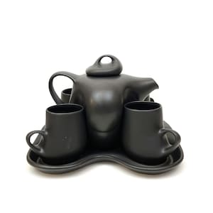 May include: A matte black ceramic tea set, including a teapot with a unique handle and lid, two matching mugs, and a tray. The set has a modern, minimalist design, perfect for serving tea. The mugs have a curved handle.