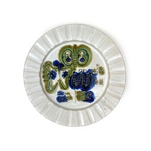 May include: White ceramic plate with a ruffled edge. The plate features a colorful, abstract design in shades of green, blue, and brown. The design appears to be a stylized floral or insect motif. The plate is round and has a slightly raised rim.