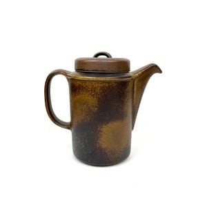 May include: A ceramic coffee pot with a lid. The pot is a dark brown color with lighter brown and tan accents. It has a handle and a spout. The lid has a small loop handle. The pot is likely vintage or antique.