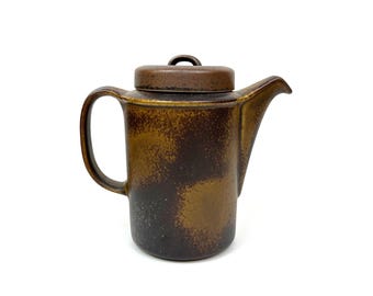 Puede incluir: Una cafetera de cerámica con tapa. La cafetera es de color marrón oscuro con acentos marrones claros y beige. Tiene un asa y un pico. La tapa tiene un pequeño asa de bucle. La cafetera es probablemente vintage o antigua.