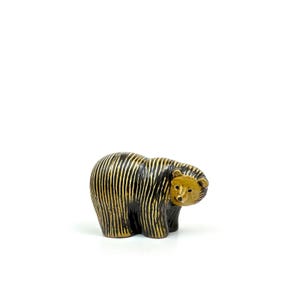 May include: A small bear figurine featuring a dark brown body with vertical tan stripes. The bear has a solid tan face with a black nose and eyes, and its head is turned slightly to the right. The surface of the figurine has a textured finish.