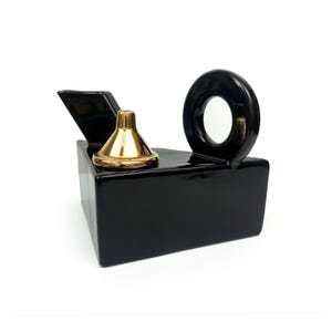May include: A glossy black ceramic teapot with a geometric, angular body. It features a gold-colored metallic conical lid, a wide angled spout, and a circular handle integrated into the top edge of the vessel, creating a distinct, modern silhouette.