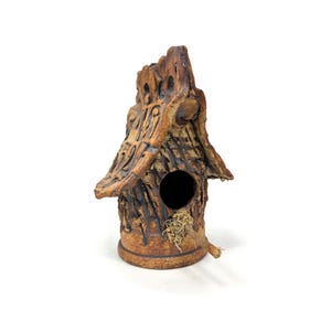 Può includere: Una casetta per uccelli in ceramica con una finitura rustica e testurizzata. La casa ha una piccola apertura per gli uccelli per entrare e un posatoio sul lato. La casa è dipinta in tonalità di marrone e beige.