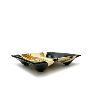 May include: A black and white ceramic dish with brown stripes. The dish has three legs and is slightly curved.