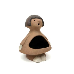May include: A brown ceramic figurine of a person with a black bob haircut and a large open mouth. The figurine has a hollow space in the front, which could be used as a small dish or trinket holder.