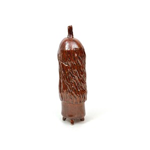 May include: A brown ceramic vase with a textured, bark-like design. The vase has a rounded top and a narrow, cylindrical body. It stands on three small feet and has a small, pointed top. The vase is glazed with a glossy finish.