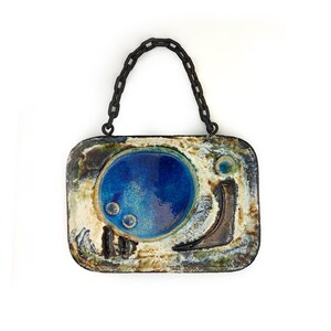 May include: Rectangular ceramic wall hanging with a dark blue chain handle. The ceramic piece features a central blue circle with two smaller circles, surrounded by textured beige, brown, and green areas. The piece has a unique, artistic design.