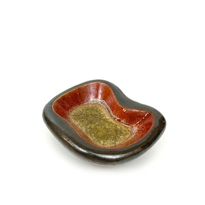 Puede incluir: Un pequeño cuenco de cerámica hecho a mano con una forma orgánica única. El cuenco presenta un exterior marrón oscuro, un interior rojo vibrante y un centro dorado. Esta pieza decorativa es ideal para guardar objetos pequeños.