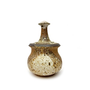 以下が含まれることがあります： 蓋付きの小さく、茶色と白の陶器の壺。壺は斑点のある釉薬と丸みを帯びた形をしています。蓋の上には小さなつまみがあります。