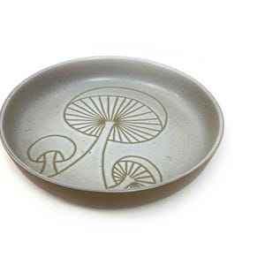 May include: A round, ceramic dish with a light gray interior and a brown rim. The dish features a detailed illustration of three mushrooms in a light brown color. The design is centered on the dish, creating a nature-inspired aesthetic.