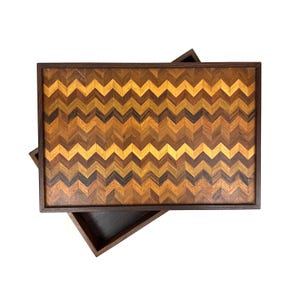 May include: Rectangular wooden tray with a dark brown frame and a chevron pattern. The pattern is made of various shades of brown and yellow wood pieces. A second, smaller tray is partially visible beneath the main tray.