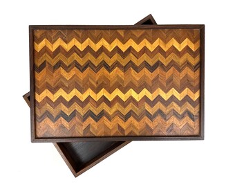 Puede incluir: Bandeja rectangular de madera con marco marrón oscuro y diseño de espiga. El diseño está hecho de varias tonalidades de madera marrón y amarilla. Una segunda bandeja, más pequeña, es parcialmente visible debajo.