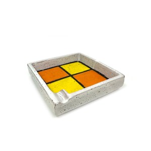 May include: Square ceramic ashtray with a speckled, off-white exterior. The interior features a vibrant design of four squares: two orange and two yellow, outlined in black. A small notch is visible on one side.