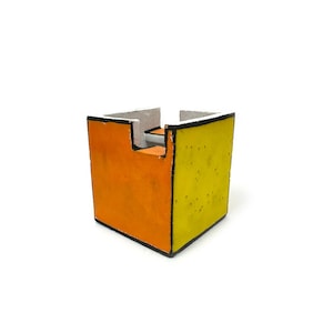 Op de afbeelding: Een kleine, vierkante, handgemaakte container met een uniek ontwerp. Eén kant is oranje, de aangrenzende kant is geel en de boven- en binnenkant zijn grijs. De randen zijn zwart omlijnd.