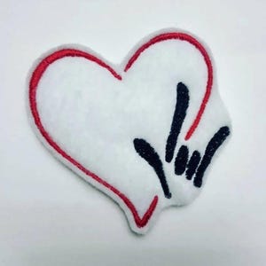May include: White heart-shaped patch with a red embroidered outline. The patch features a black hand gesture and a red accent. The patch is made of a soft, textured material.