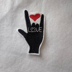 May include: Black and white embroidered patch in the shape of a hand making the "I love you" sign. A red heart is above the fingers, and the word "LOVE" is embroidered in white.