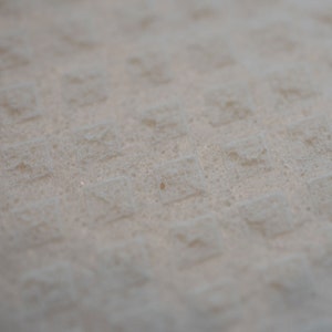 May include: Close-up of a textured, off-white fabric. The surface is covered in a repeating pattern of small, raised squares, creating a subtle grid-like design. The fabric appears soft and slightly blurred, suggesting a focus on texture.