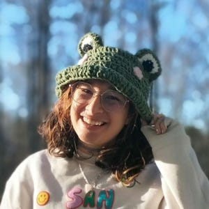 May include: A green crocheted frog hat with large eyes and pink accents. The hat is worn by a person wearing glasses and a white sweatshirt with the word "SAN" in colorful letters. The background is blurred with sunlight.