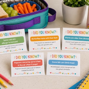 May include: An open teal and purple lunch bag contains a sandwich, grapes, and carrots. Five cards with the text "DID YOU KNOW?" display facts about animals and food. Colourful pencils and a water bottle are also visible. The cards are in English.